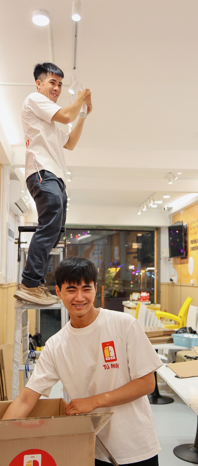 Một nhân viên trên thang tháo đèn trần, một người dưới giữ thùng
